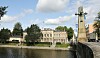Historiallinen rakennus joen varrella, edessä puistoa ja oikealla korkea muistomerkki.