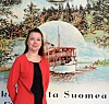 Maarika Kasonen seisoo punaisessa jakussa Suomi-aiheisen taustakuvan edessä.