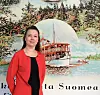 Maarika Kasonen seisoo punaisessa jakussa Suomi-aiheisen taustakuvan edessä.